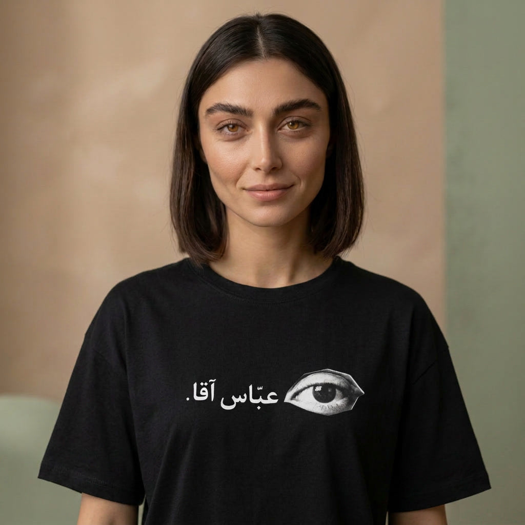 A woman with shoulder-length dark brown hair wearing a black t-shirt with a white Arabic text graphic and an eye illustration. The background is a soft-focus beige and green wall.