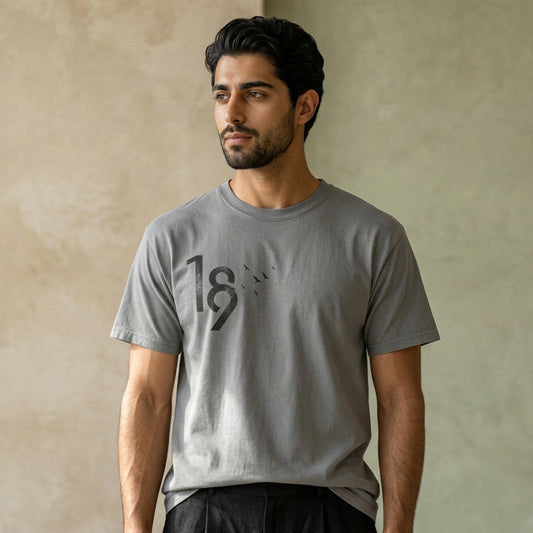 A man wearing a light gray crewneck t-shirt with a black graphic print of the number 18, paired with dark charcoal pants, standing against a textured beige wall.
