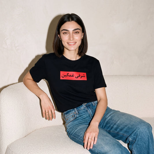 A woman with dark shoulder-length hair sits on a textured white armchair, wearing a black crewneck t-shirt with a red rectangular graphic containing black Arabic text, paired with blue jeans.